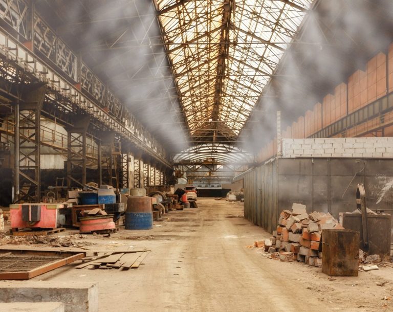 Innenansicht einer alten, verlassenen Fabrik mit Rohren und Maschinen in staubiger Umgebung.